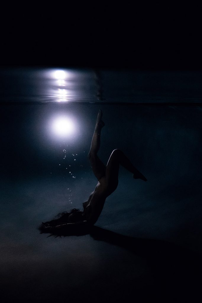 A woman laying at the bottom of a pool at night with her legs pointed to the surface during her Florida underwater photography session.