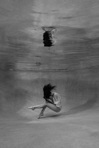 A woman sits on the floor of a pool, holding her head in her hands, under her own reflection.