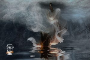 A woman underwater in a white robe floats upside to the surface with her face breaking through, during her underwater photography photoshoot.